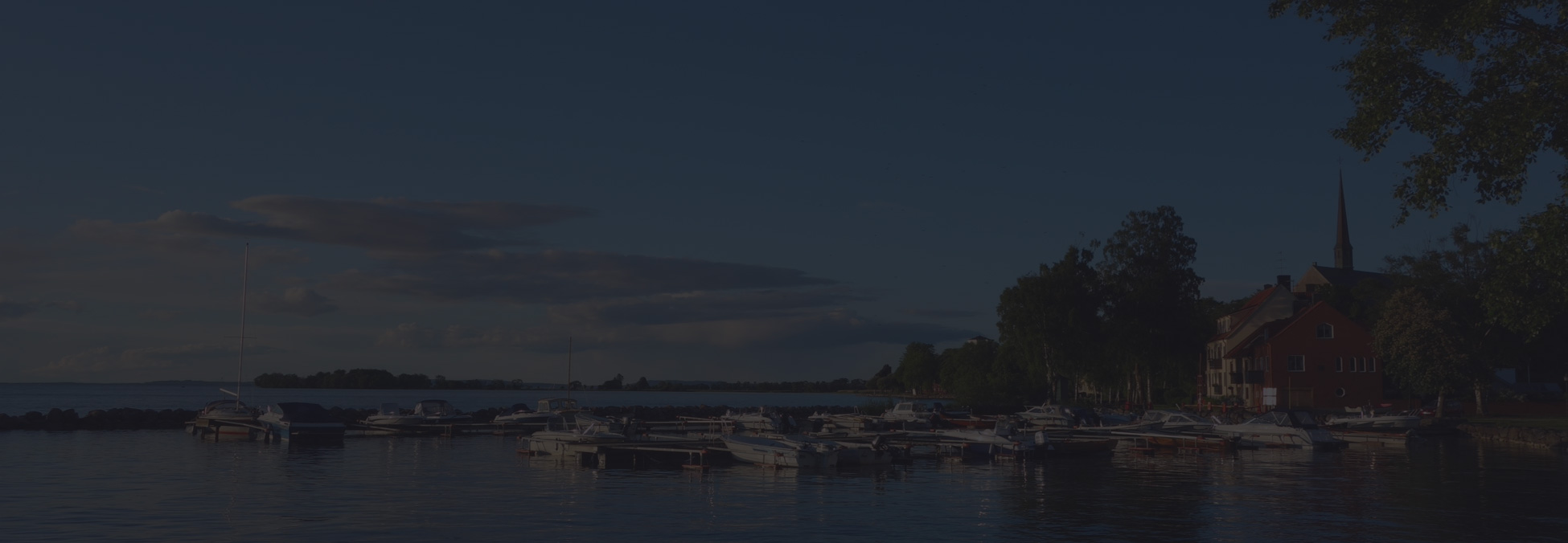 A calm evening waterfront scene with small docked boats, trees, and a distant church spire under a blue dusky sky; tranquil lakeside mood.