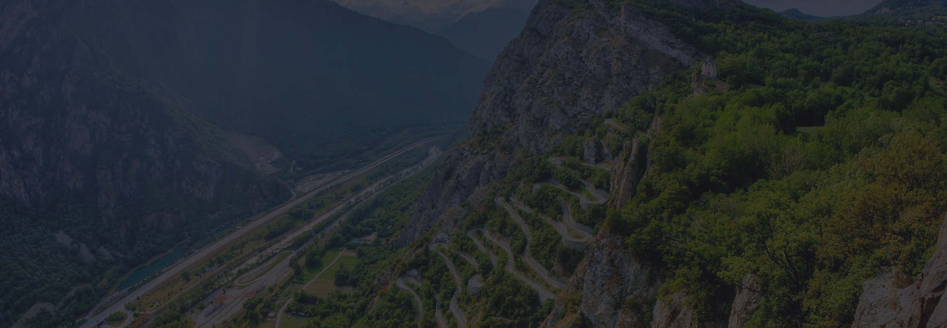 A winding mountain road cut through rocky cliffs with dense green forest, dramatic and expansive mood